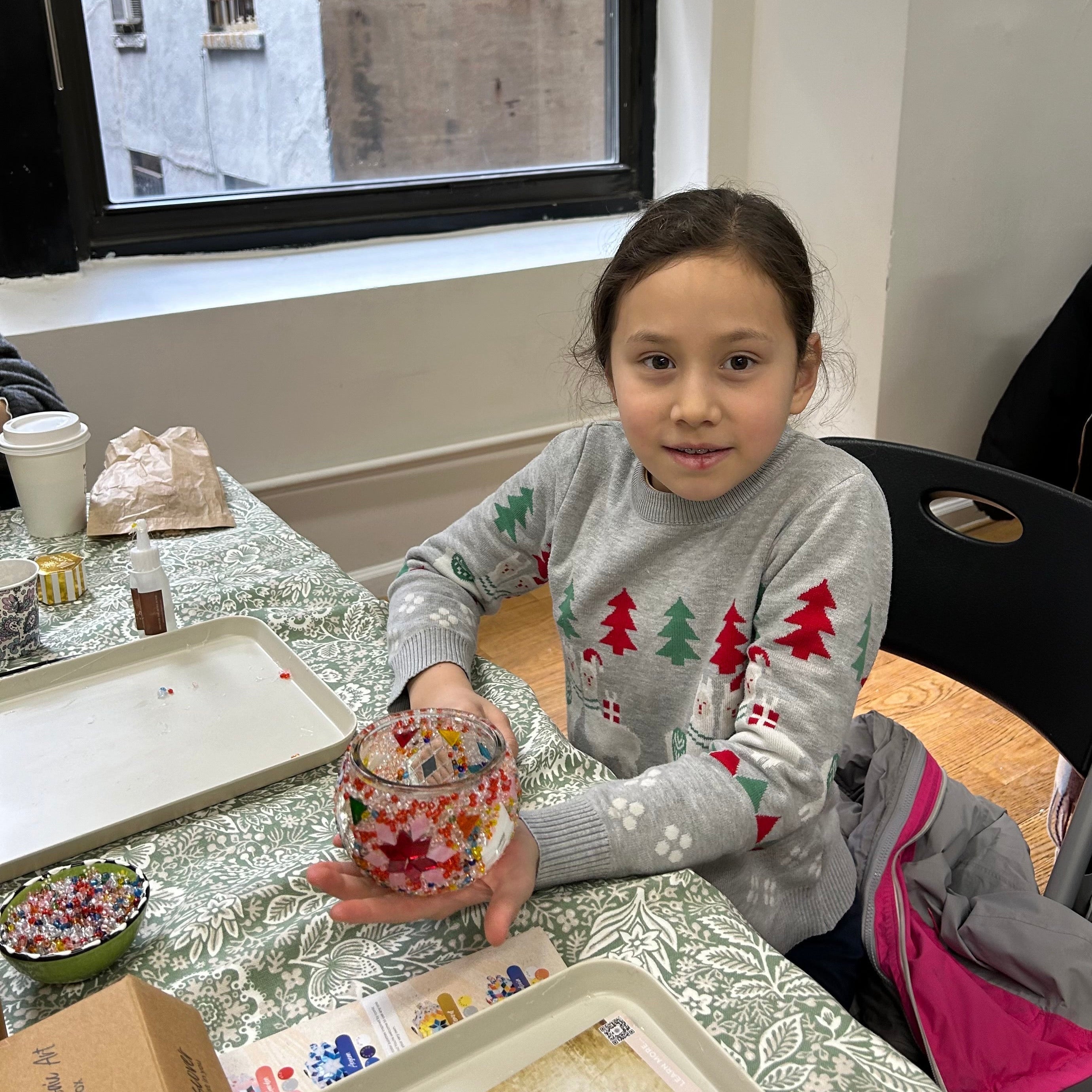 Child in Christmas sweater holding a decorated mosaic glass jar at a craft table