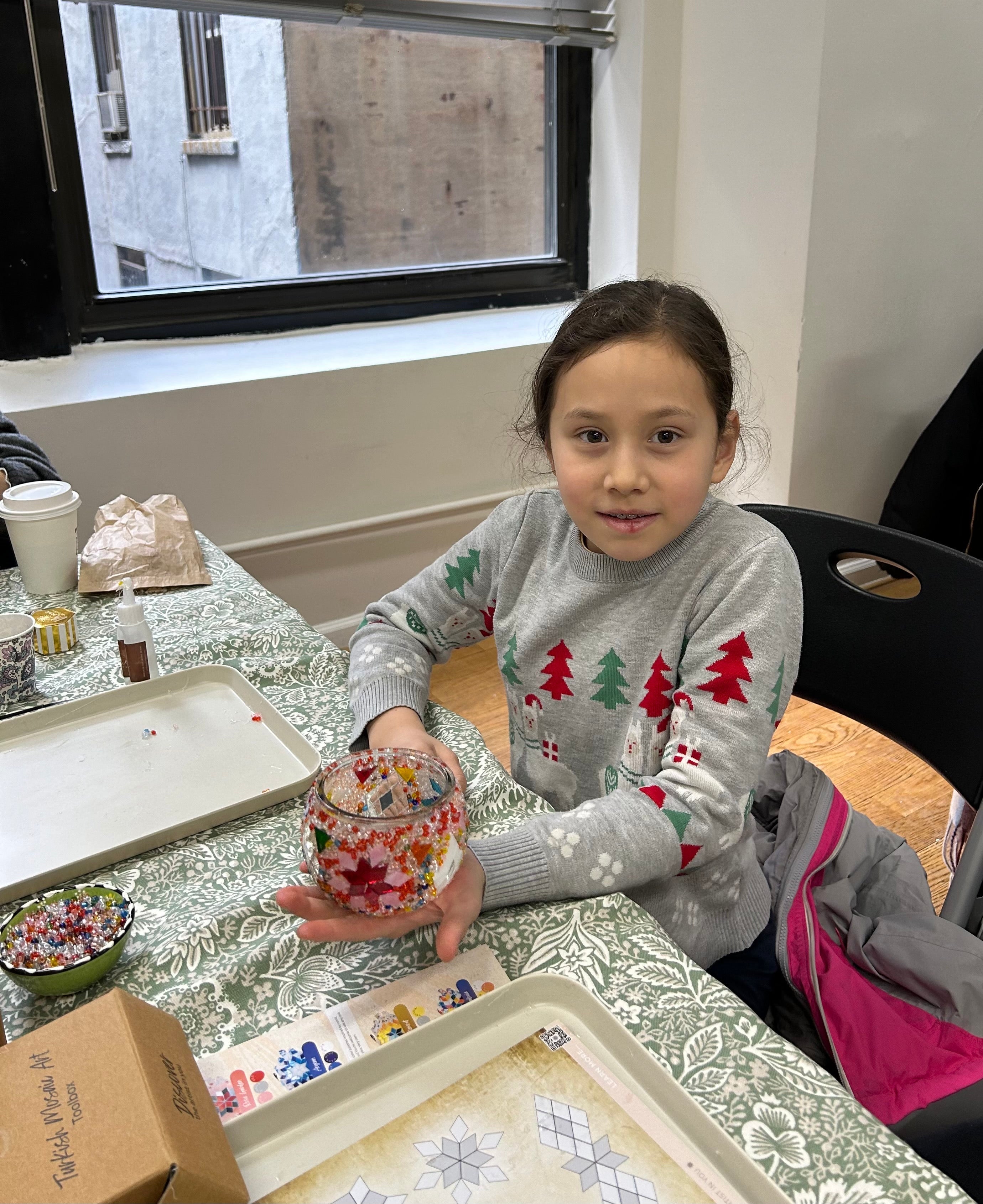 Child in Christmas sweater holding a decorated mosaic glass jar at a craft table