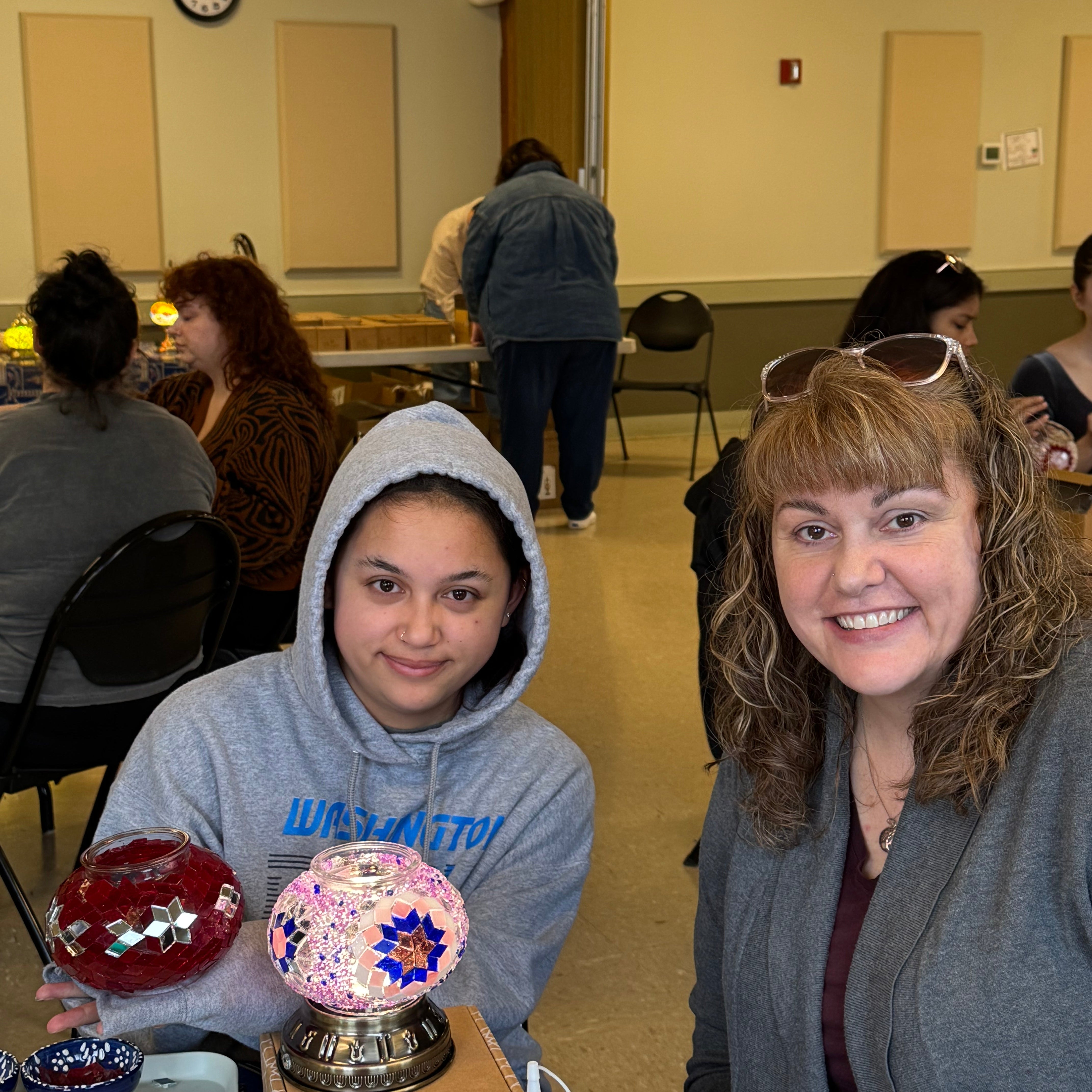 Two women smiling indoors at a craft table with mosaic glass lamps and art supplies
