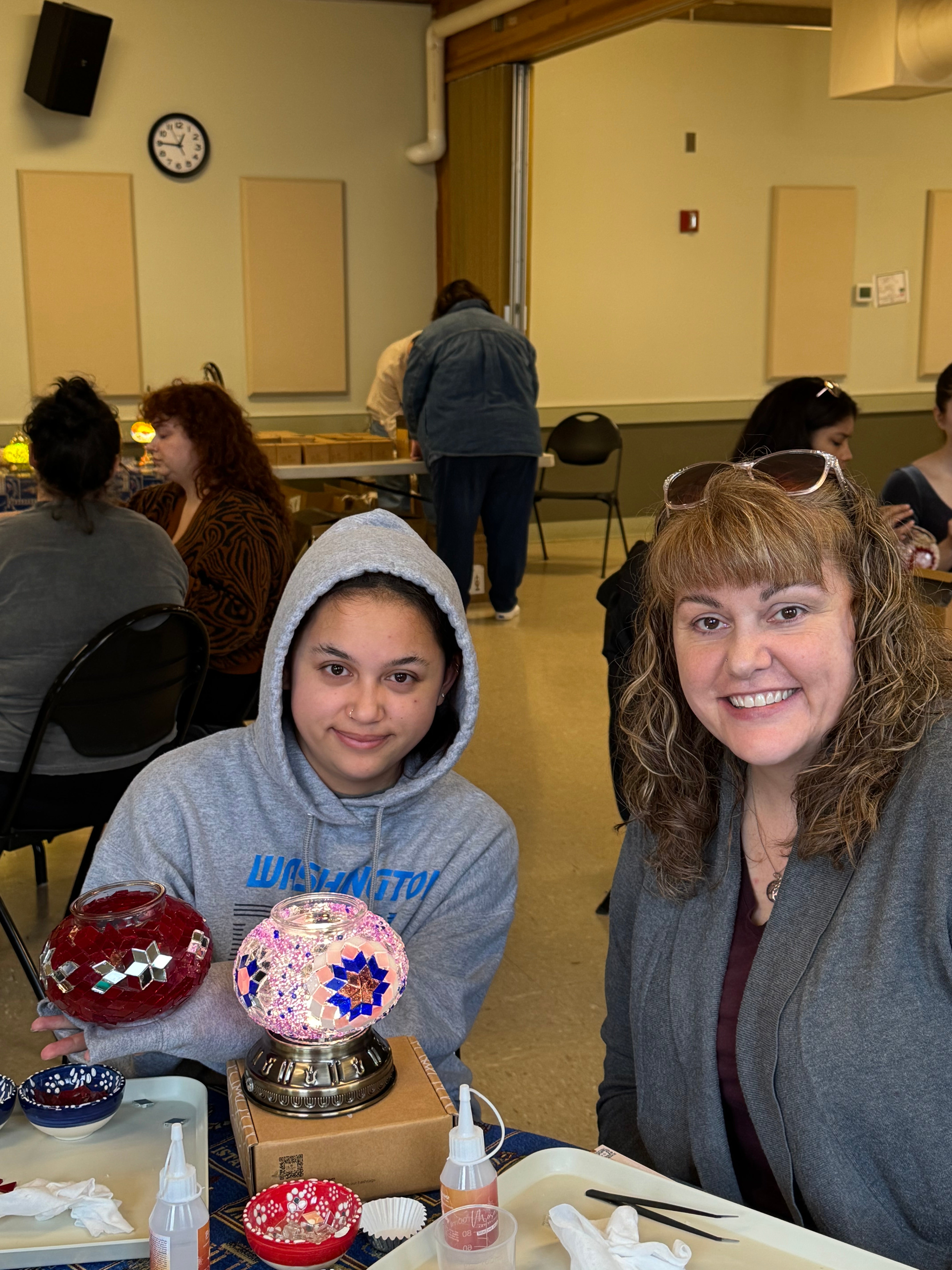 Two women smiling indoors at a craft table with mosaic glass lamps and art supplies