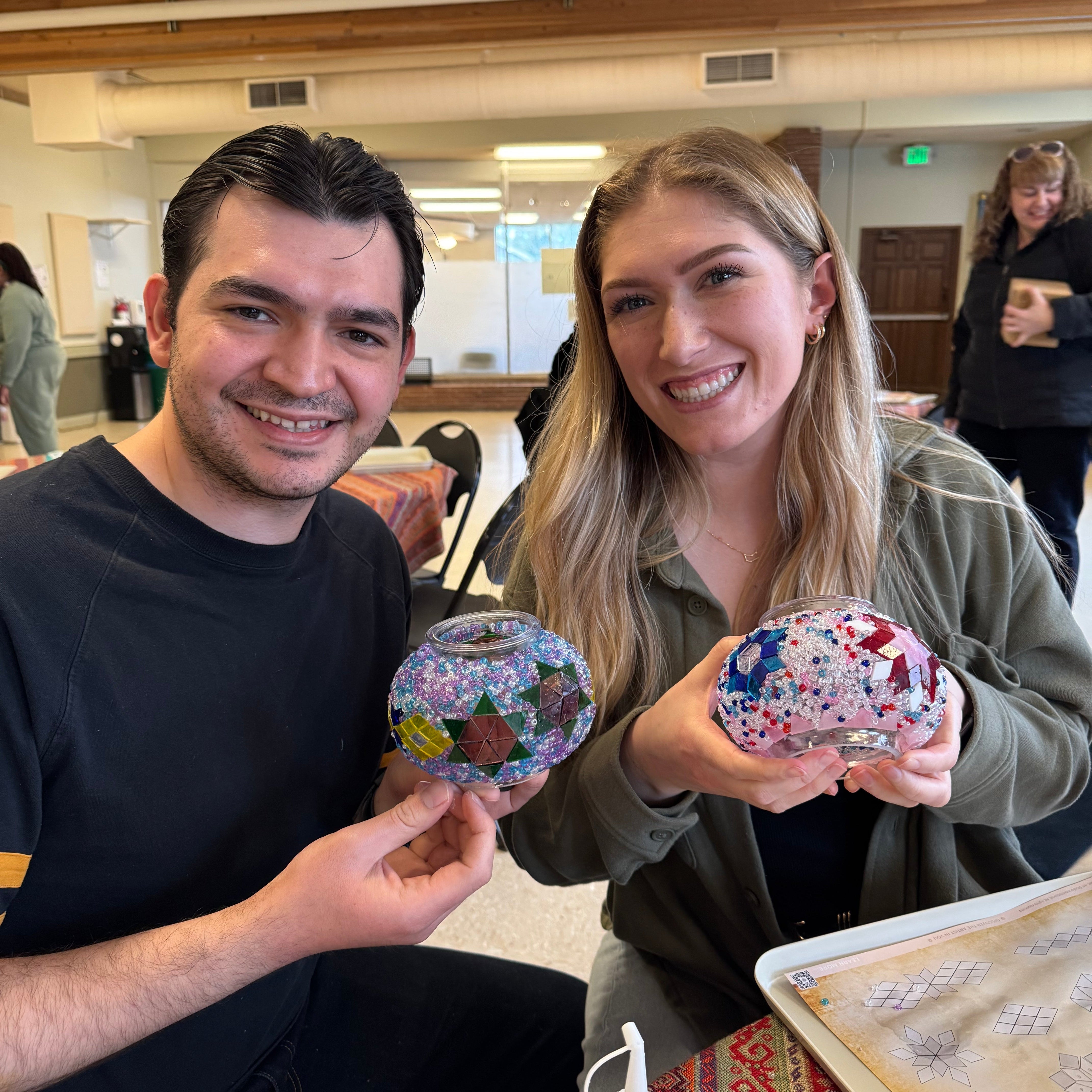 Smiling man and woman holding colorful mosaic glass bowls in a bright craft workshop