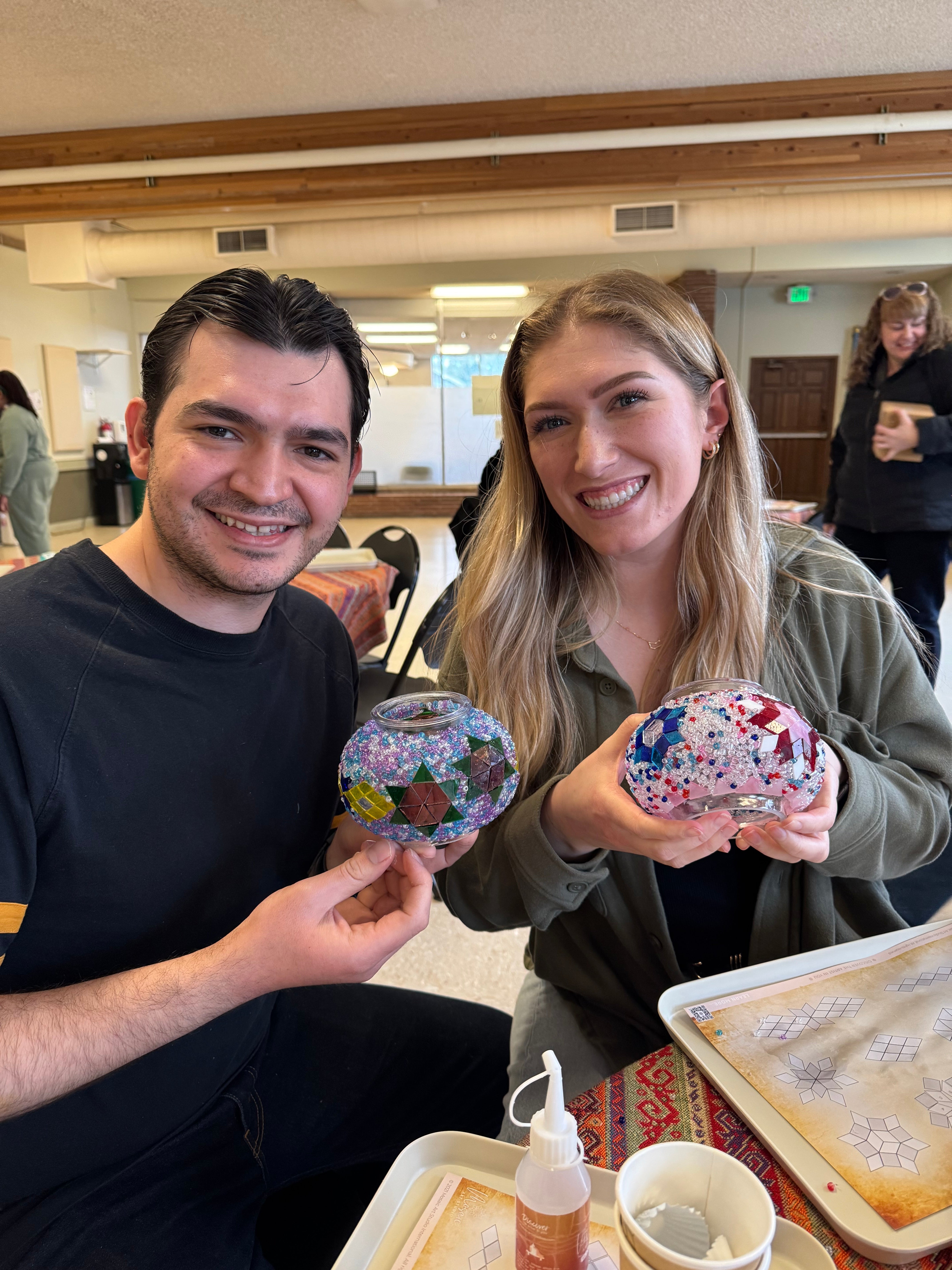 Smiling man and woman holding colorful mosaic glass bowls in a bright craft workshop