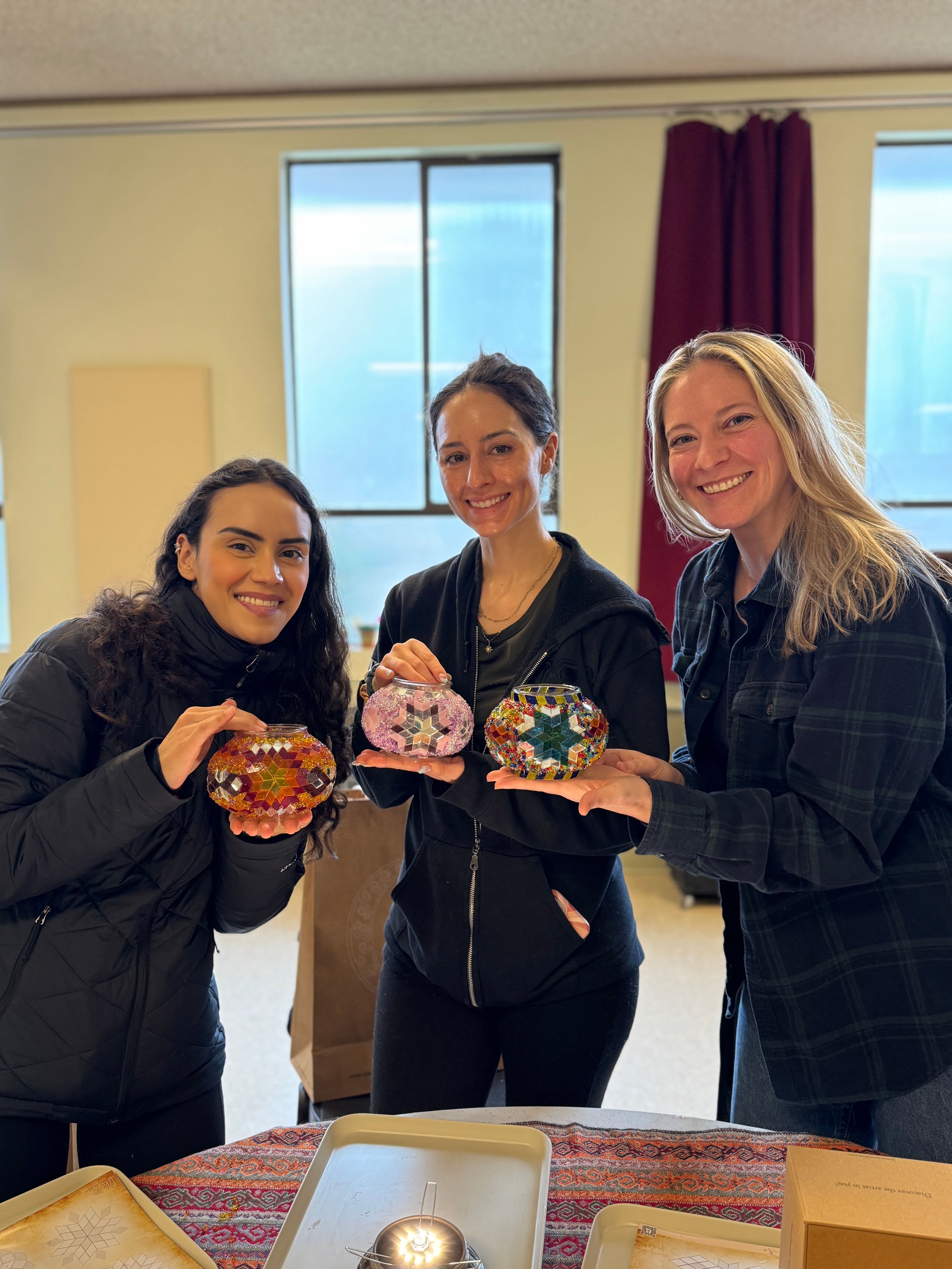 Three women smiling indoors holding colorful mosaic glass candle holders over a patterned table