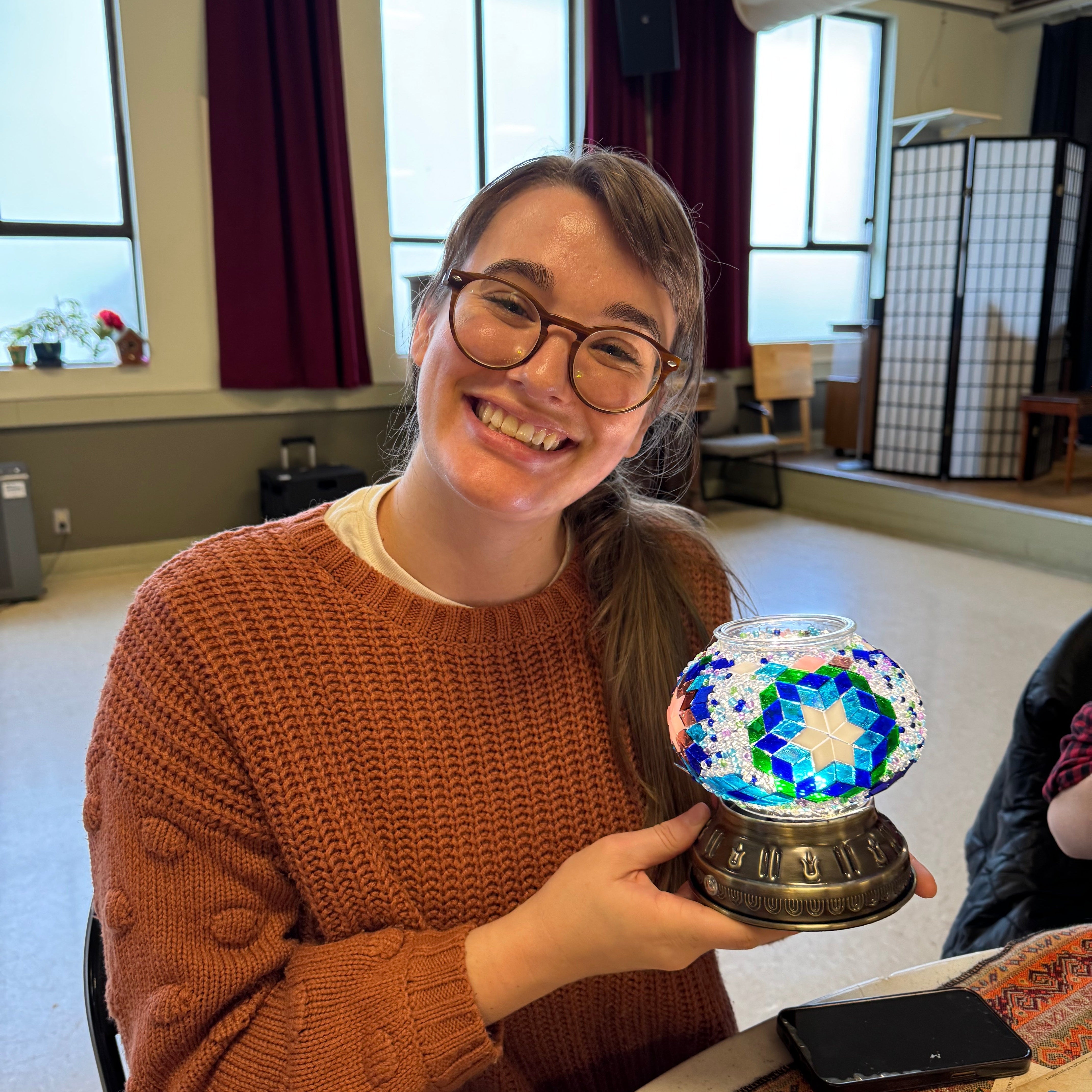 Smiling woman in brown sweater holding colorful mosaic glass lamp in cozy craft room