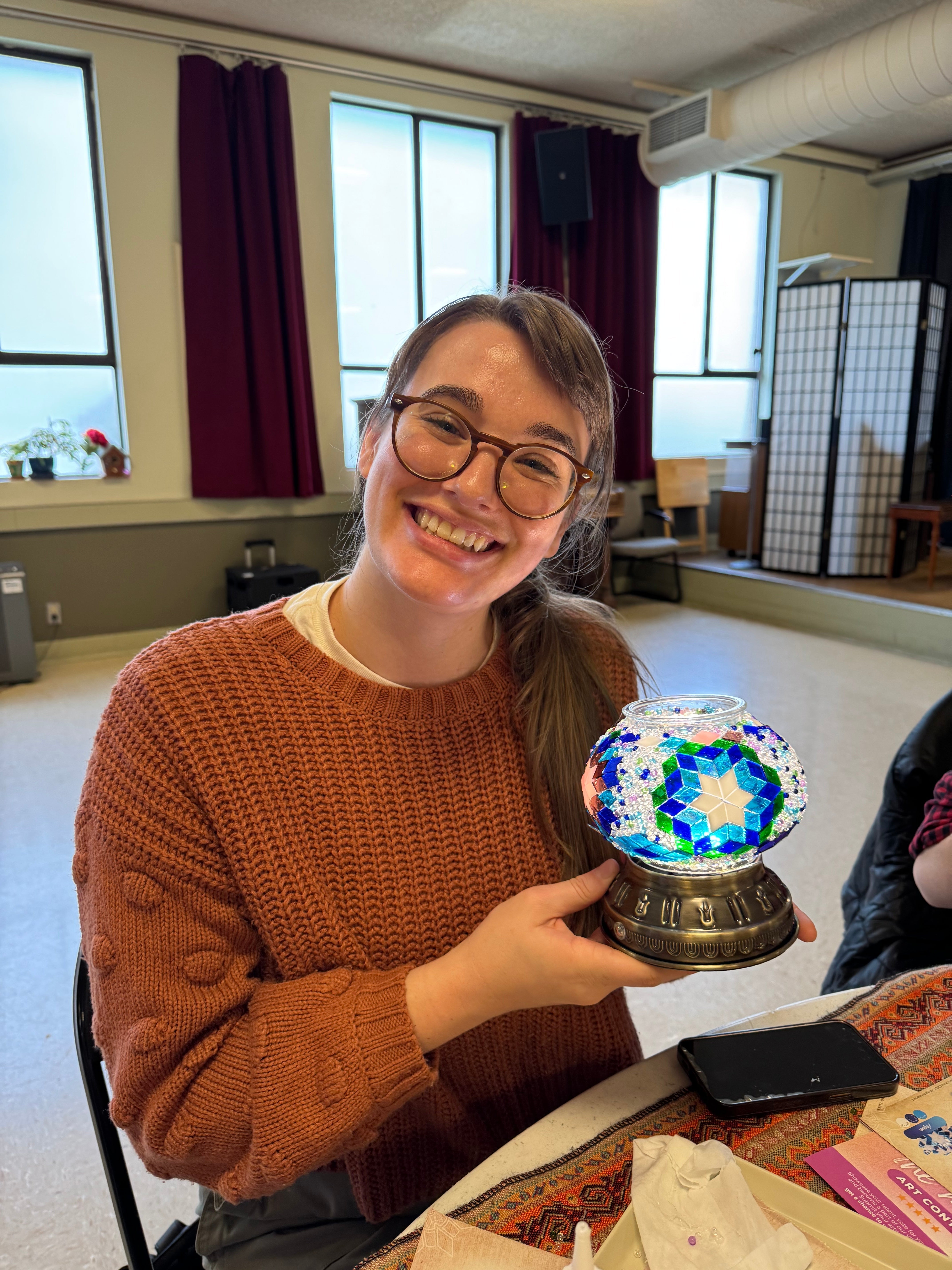 Smiling woman in brown sweater holding colorful mosaic glass lamp in cozy craft room