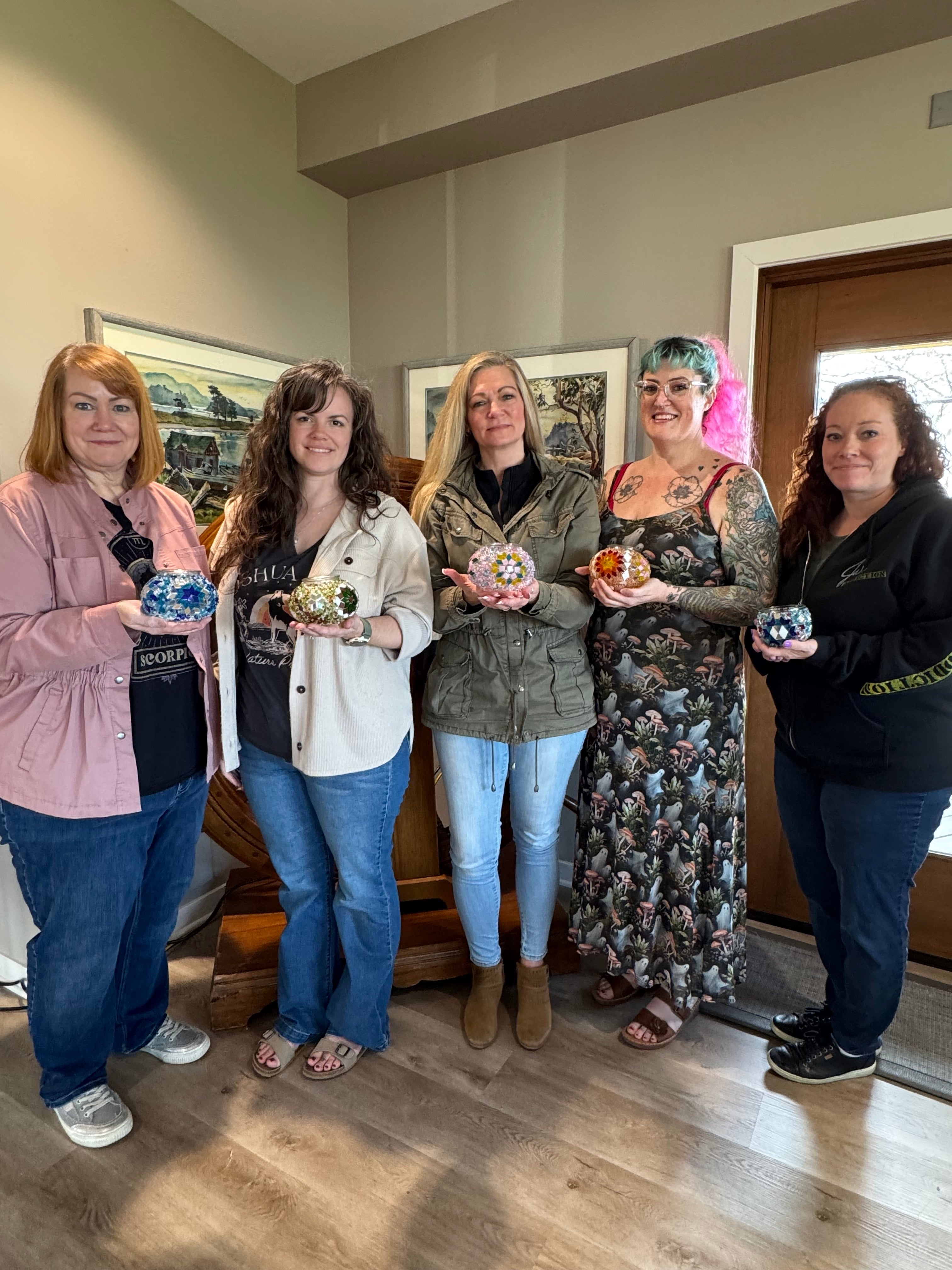 Five women standing indoors holding colorful mosaic glass candle holders, casual setting with wooden floor