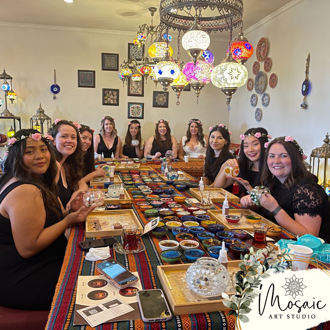 Group of women with flower crowns at a mosaic art studio table with colorful tiles and lanterns