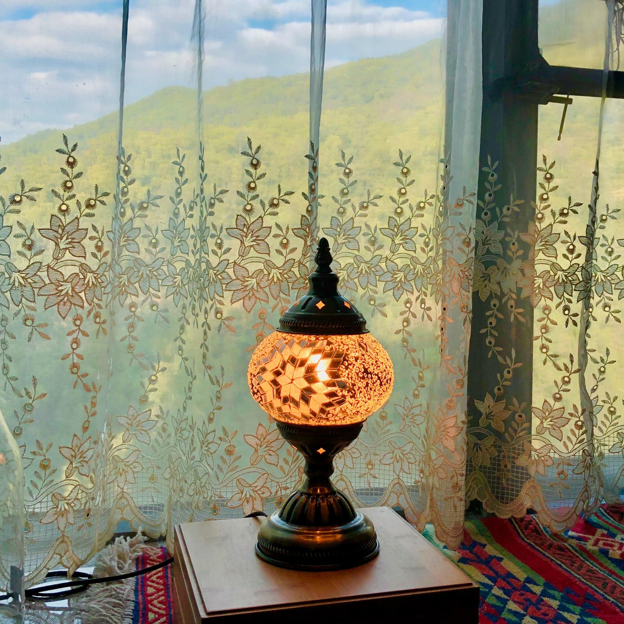 Turkish mosaic lamp with warm glowing lava design on wooden table by floral lace curtains and window