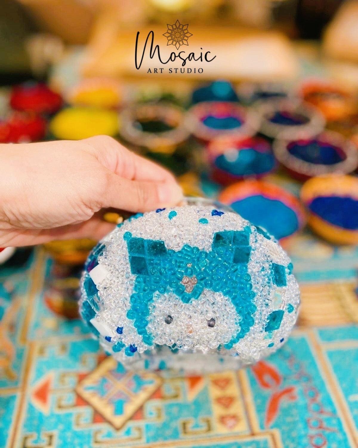 Hand holding a beaded turquoise and white mosaic art piece on a colorful patterned surface