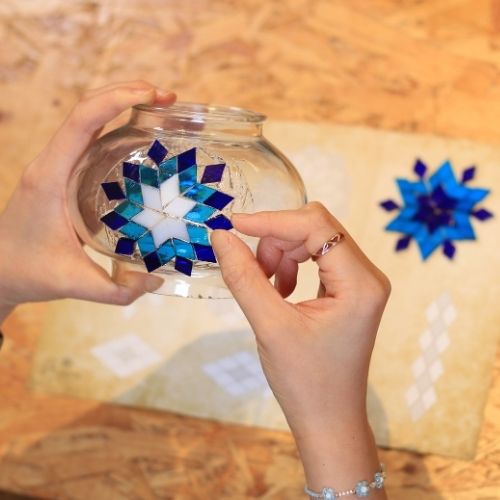 Hands applying blue and white mosaic star pattern to clear glass bowl on wooden table