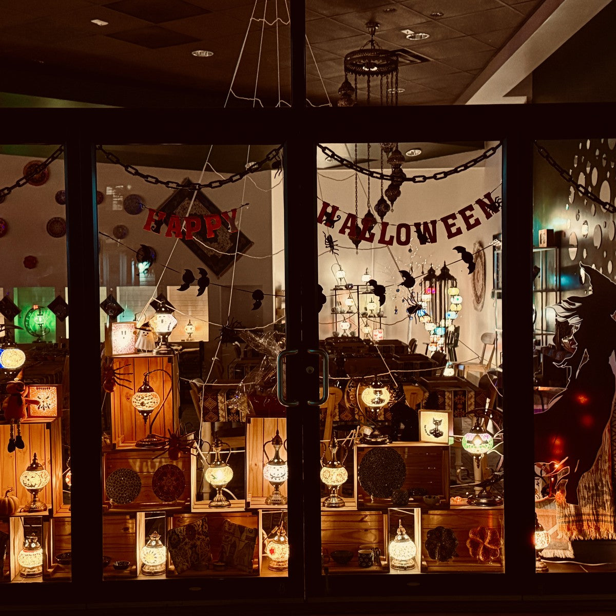 Mosaic Art Studio Halloween storefront window at night filled with glowing mosaic lamps and paper garlands
