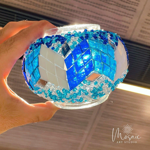 Hand holding a blue and white Turkish mosaic lamp with glass pieces and bead decoration under ceiling lights