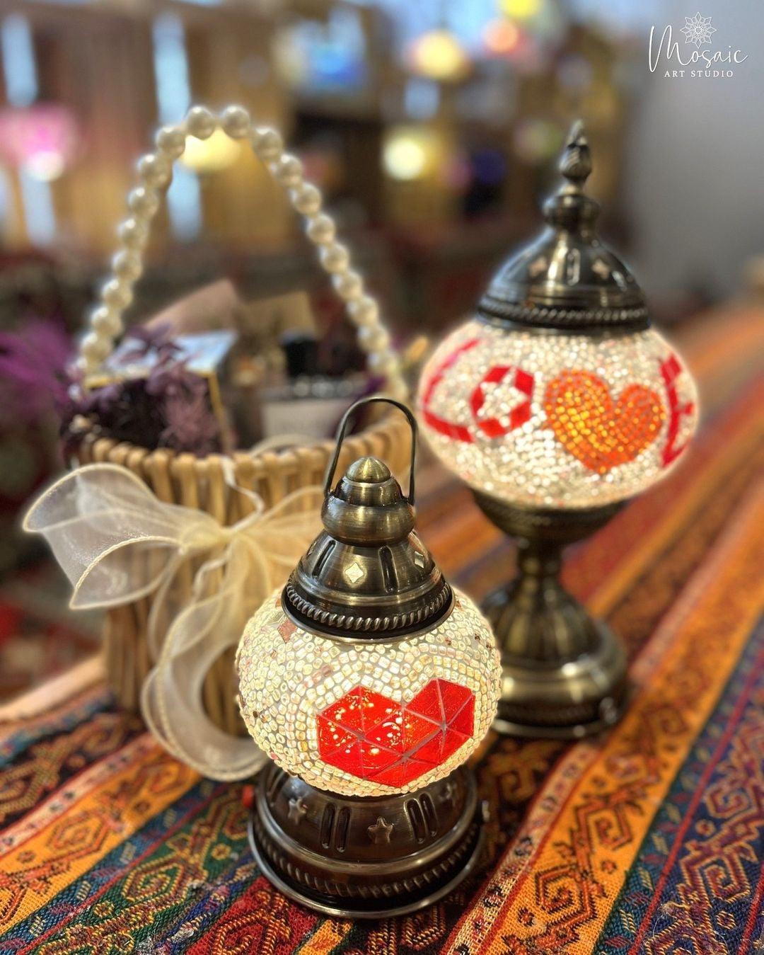 Two illuminated Turkish mosaic lamps with heart designs on a colorful patterned fabric with a decorated basket in the background