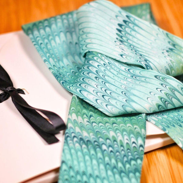 Close-up of teal patterned silk scarf with peacock feather design on a white gift box with black ribbon