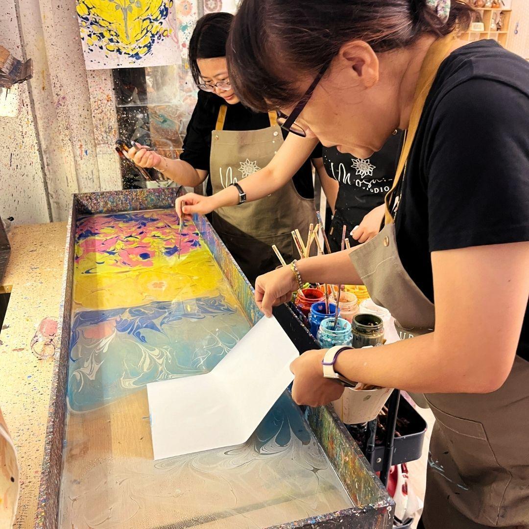 Two women in aprons creating colorful marbled art on water surface using paper and paint in a studio