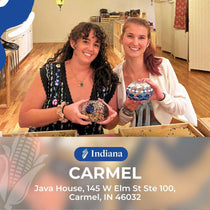Two smiling women holding handmade mosaic lamps inside a workshop with wooden floors and art displays