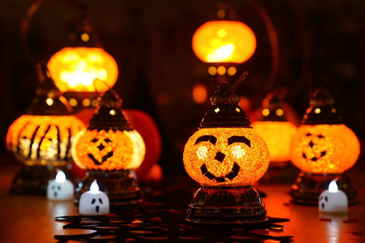 Jack-o'-lantern mosaic lamps made of orange glass glowing on a table beside small ghost candles
