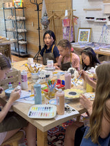Students painting ceramic pieces at a shared table stocked with paints and brushes