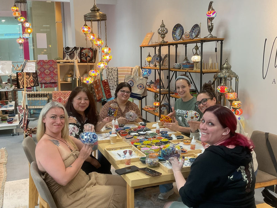 Guests at a mosaic lamp workshop table inside the Voorhees, NJ studio, with lamps and ceramics on the shelves