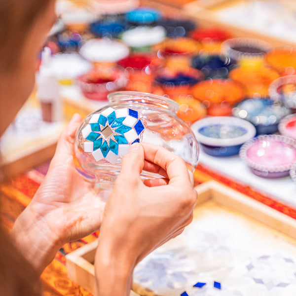 Hands placing blue and white glass tiles into a star pattern on a clear glass lamp globe
