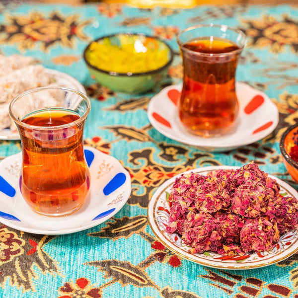 Two glasses of Turkish tea on colorful saucers with plates of rose petal sweets and nuts on a patterned tablecloth