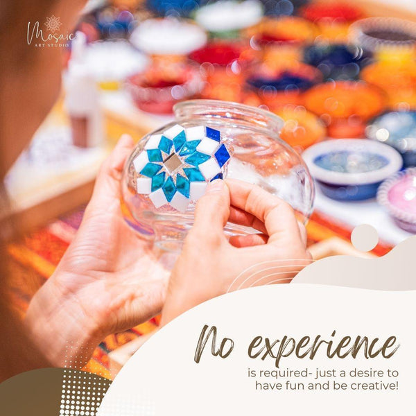 Hands placing blue and white mosaic tiles on a clear glass jar in a colorful mosaic art studio workshop