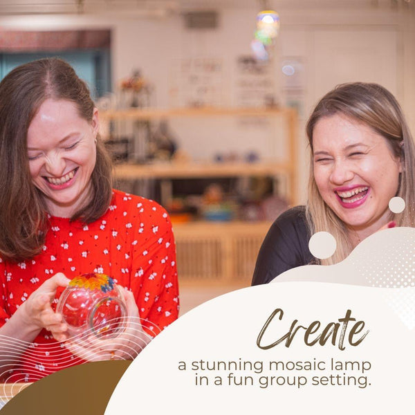 Two women laughing together while crafting a mosaic lamp in a cozy indoor workshop