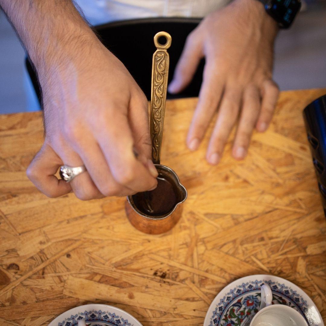 Hands stirring Turkish coffee in a traditional copper cezve on a wooden table with coffee cups