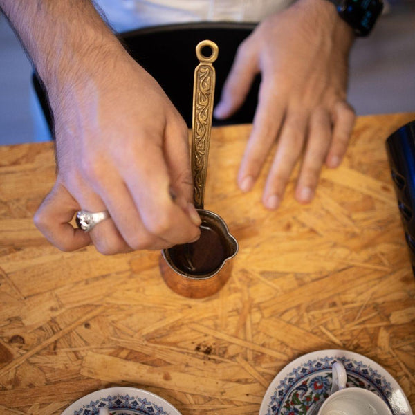 Hands stirring Turkish coffee in a traditional copper cezve on a wooden table with coffee cups