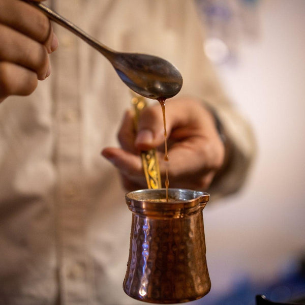 Pouring thick Turkish coffee from a spoon into a copper cezve pot held by a person in beige shirt