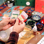 Hands holding packets of green glass beads and red handcut mosaics for DIY mosaic candle holder kit