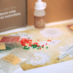 Christmas mosaic candle holder DIY kit with colored beads, glue bottle, and tweezers on patterned paper