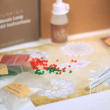 Christmas mosaic candle holder DIY kit with colored beads, glue bottle, and tweezers on patterned paper